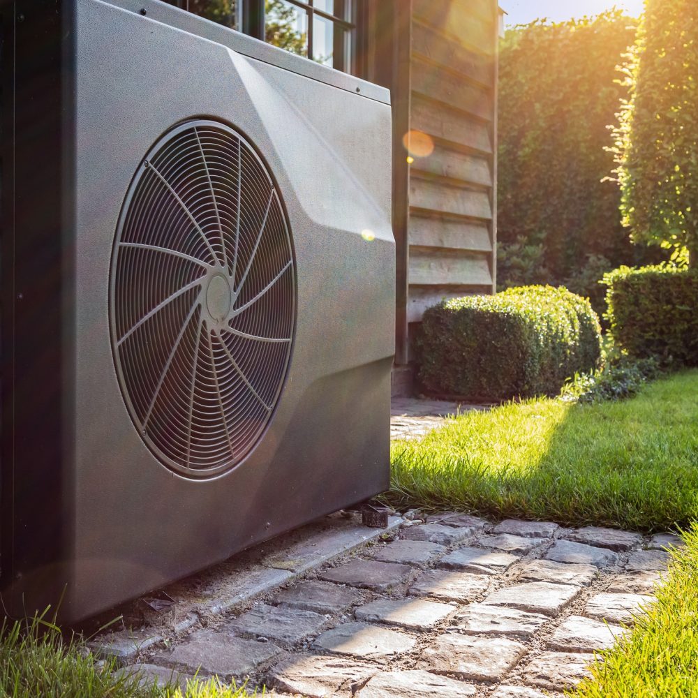 Close-up of black full inverter heat pump outside in the garden, near wooden pool house on a sunny day. Lens flare on the image.